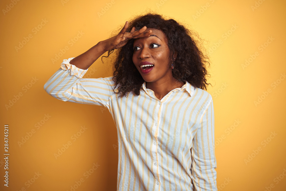 African american woman wearing striped shirt standing over isolated yellow background very happy and smiling looking far away with hand over head. Searching concept. African american woman wearing striped shirt standing over isolated yellow background very happy and smiling looking far away with hand over head. Searching concept.