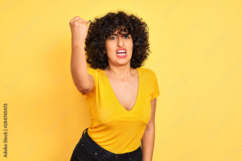 Young arab woman with curly hair wearing t-shirt standing over isolated yellow background angry and mad raising fist frustrated and furious while shouting with anger. Rage and aggressive concept. Young arab woman with curly hair wearing t-shirt standing over isolated yellow background angry and mad raising fist frustrated and furious while shouting with anger. Rage and aggressive concept.