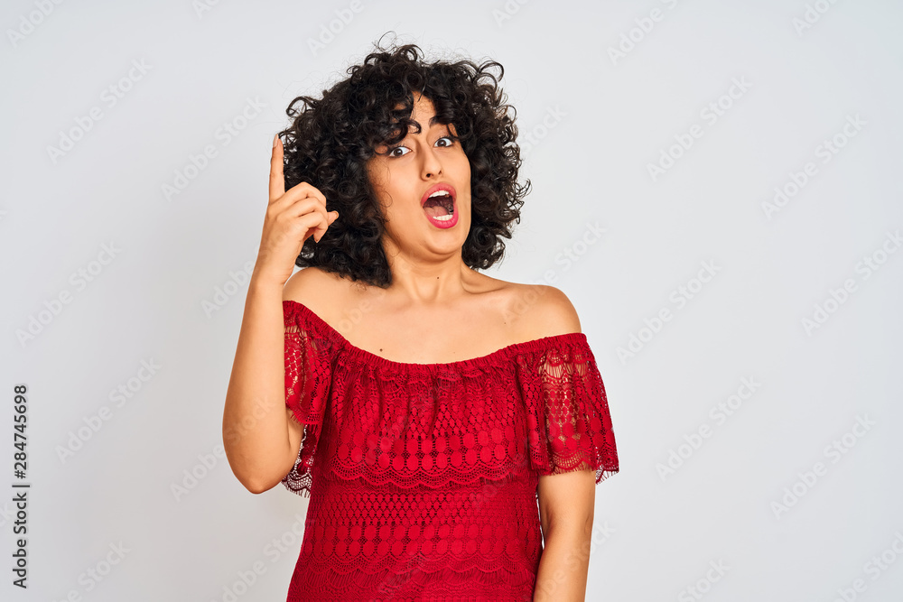 Young arab woman with curly hair wearing red dress standing over isolated white background pointing finger up with successful idea. Exited and happy. Number one. Young arab woman with curly hair wearing red dress standing over isolated white background pointing finger up with successful idea. Exited and happy. Number one.