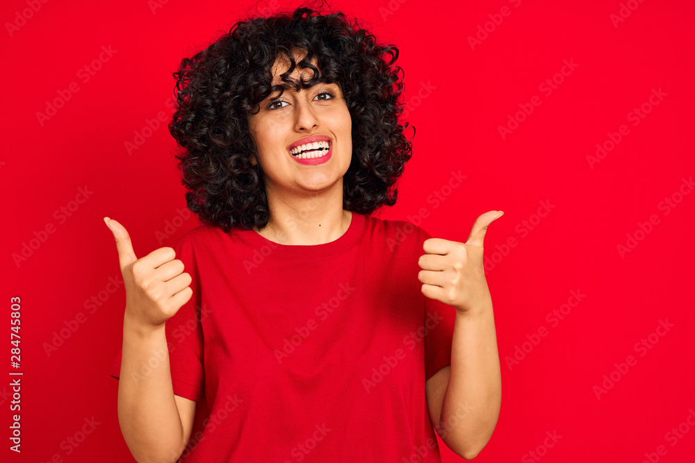 Young arab woman with curly hair wearing casual t-shirt over isolated red background success sign doing positive gesture with hand, thumbs up smiling and happy. Cheerful expression and winner gesture. Young arab woman with curly hair wearing casual t-shirt over isolated red background success sign doing positive gesture with hand, thumbs up smiling and happy. Cheerful expression and winner gesture.