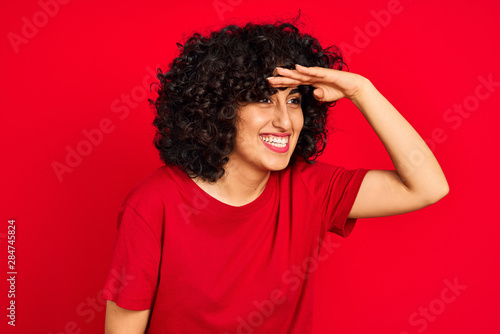 Young arab woman with curly hair wearing casual t-shirt over isolated red background very happy and smiling looking far away with hand over head. Searching concept.