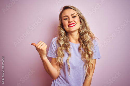 Young beautiful woman wearing purple t-shirt standing over pink isolated background smiling with happy face looking and pointing to the side with thumb up.