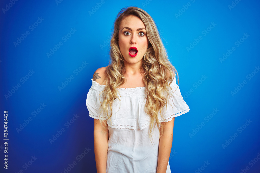 Young beautiful woman wearing white dress standing over blue isolated background afraid and shocked with surprise expression, fear and excited face. Young beautiful woman wearing white dress standing over blue isolated background afraid and shocked with surprise expression, fear and excited face.