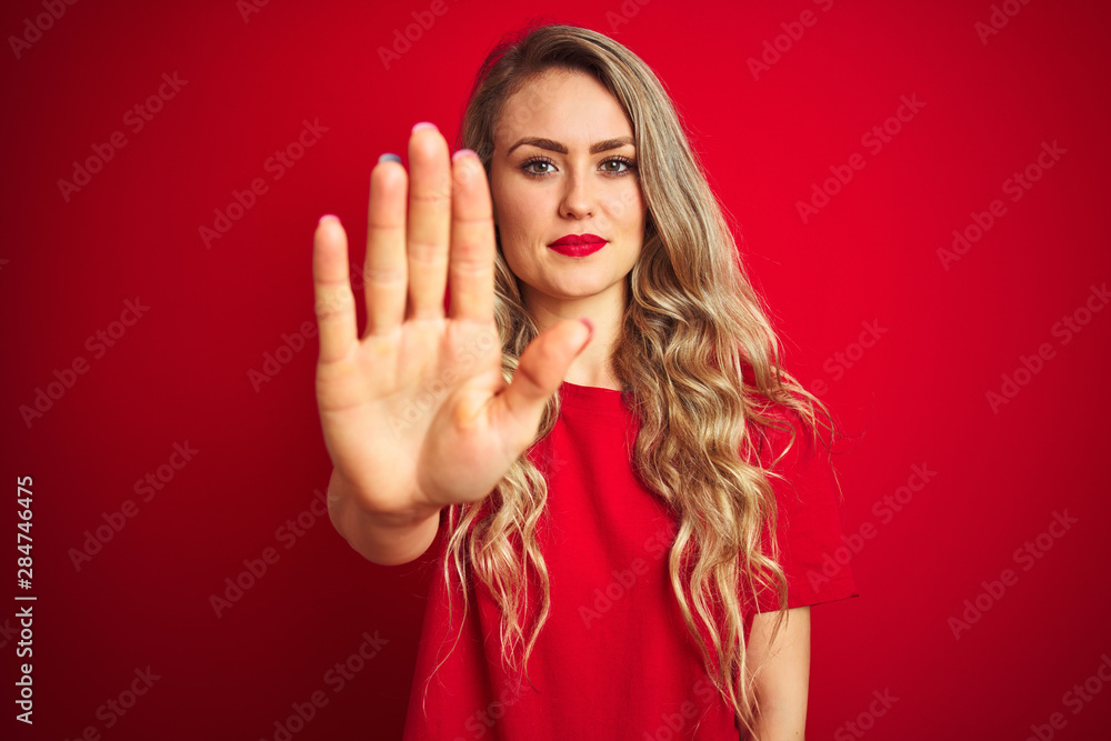 Young beautiful woman wearing basic t-shirt standing over red isolated background doing stop sing with palm of the hand. Warning expression with negative and serious gesture on the face. Young beautiful woman wearing basic t-shirt standing over red isolated background doing stop sing with palm of the hand. Warning expression with negative and serious gesture on the face.
