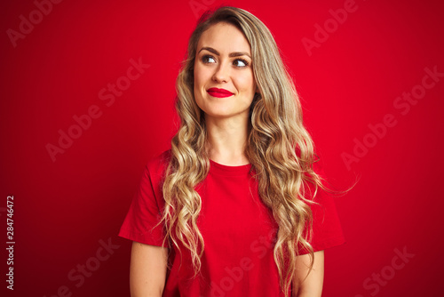 Young beautiful woman wearing basic t-shirt standing over red isolated background smiling looking to the side and staring away thinking.