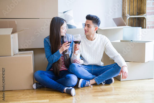 Young asian couple sitting on the floor of new apartment arround cardboard boxes, smiling drinking a glass of wine excited for moving to a new house