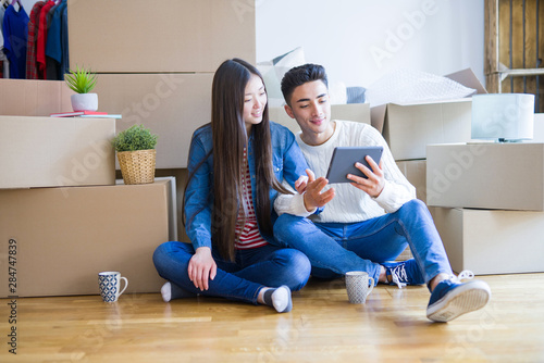Young asian couple sitting on the floor of new apartment arround cardboard boxes, using touchpad tablet and smiling at new home
