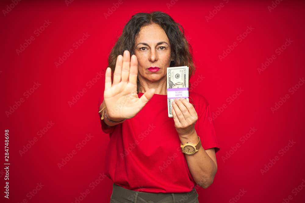 Middle age senior woman holding bunch of dollars over red isolated background with open hand doing stop sign with serious and confident expression, defense gesture Middle age senior woman holding bunch of dollars over red isolated background with open hand doing stop sign with serious and confident expression, defense gesture