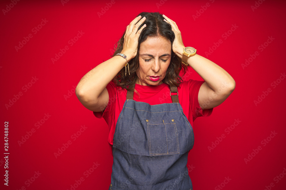 Middle age senior woman wearing apron uniform over red isolated background suffering from headache desperate and stressed because pain and migraine. Hands on head. Middle age senior woman wearing apron uniform over red isolated background suffering from headache desperate and stressed because pain and migraine. Hands on head.