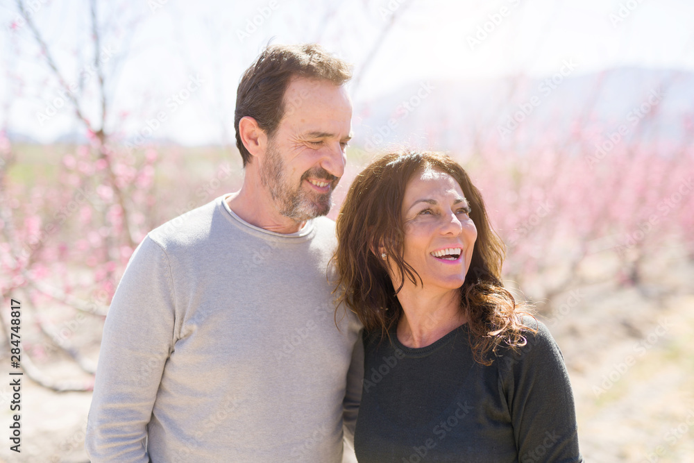 Beautiful middle age senior couple smiling in love at romantic garden of peach trees with pink petals on a sunny day of spring Beautiful middle age senior couple smiling in love at romantic garden of peach trees with pink petals on a sunny day of spring