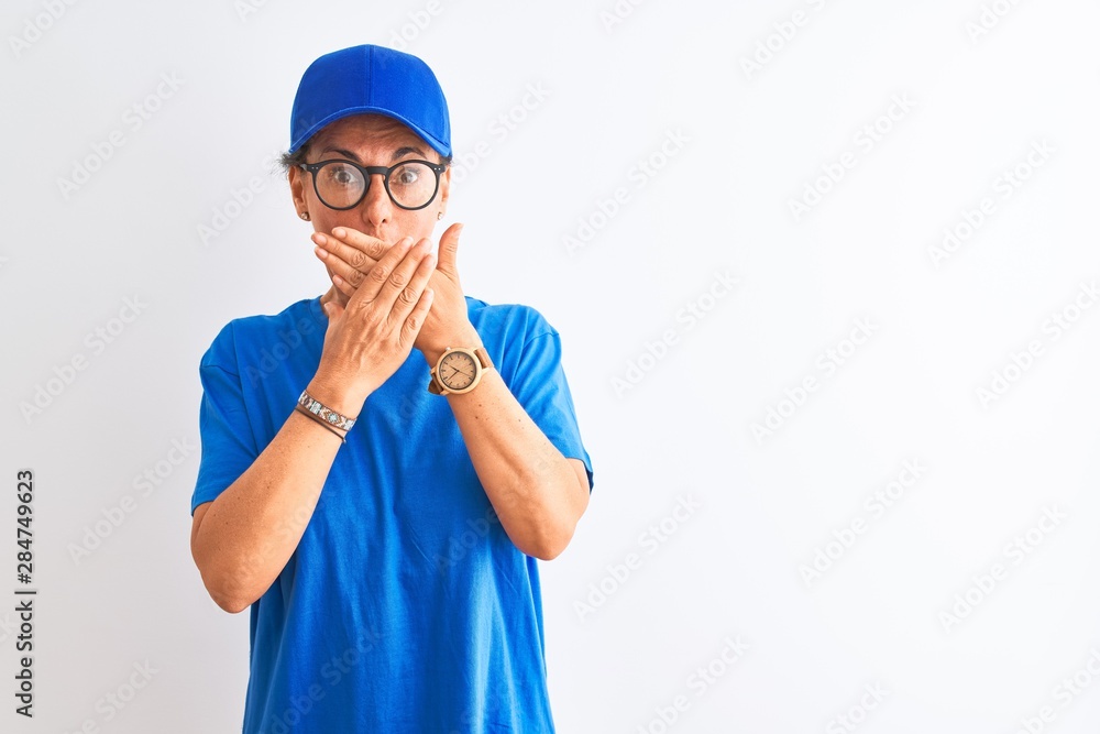 Senior deliverywoman wearing cap and glasses standing over isolated white background shocked covering mouth with hands for mistake. Secret concept.