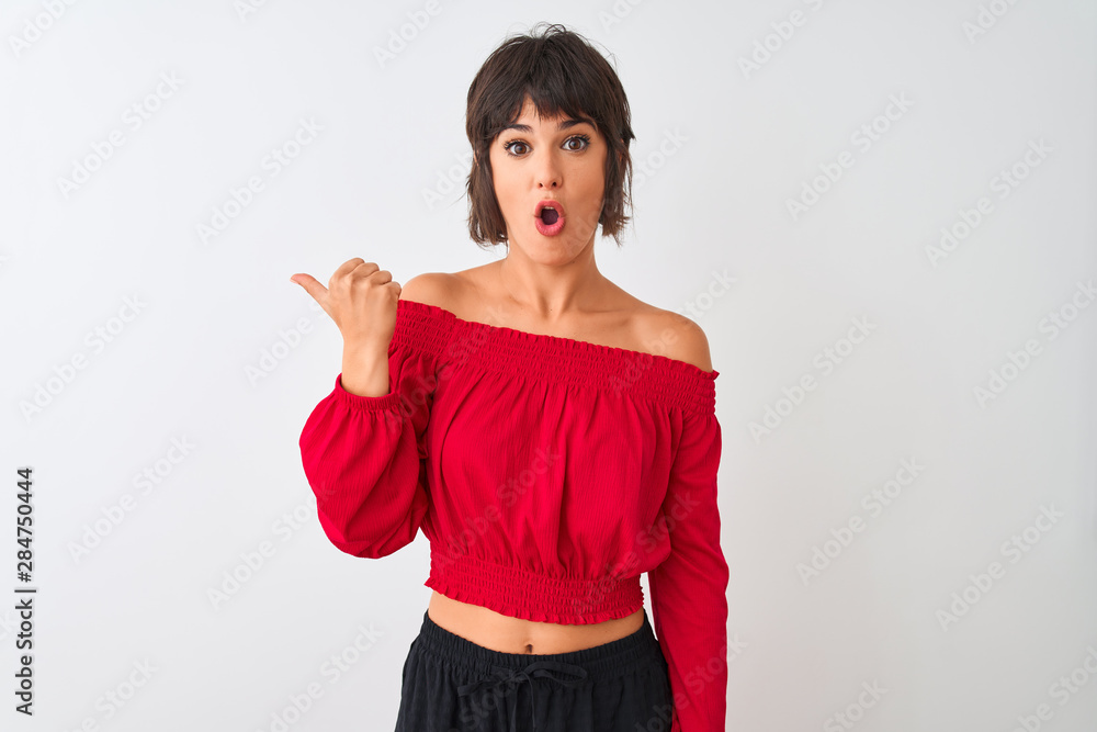 Young beautiful woman wearing red summer t-shirt standing over isolated white background Surprised pointing with hand finger to the side, open mouth amazed expression.