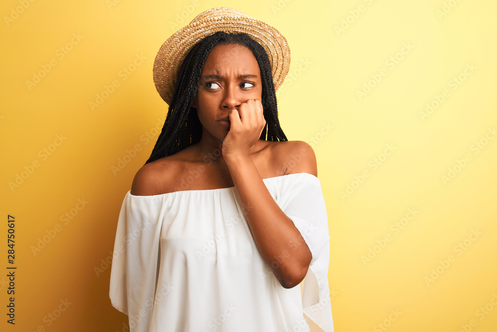 Young african american woman wearing white t-shirt and hat over isolated yellow background looking stressed and nervous with hands on mouth biting nails. Anxiety problem. Young african american woman wearing white t-shirt and hat over isolated yellow background looking stressed and nervous with hands on mouth biting nails. Anxiety problem.