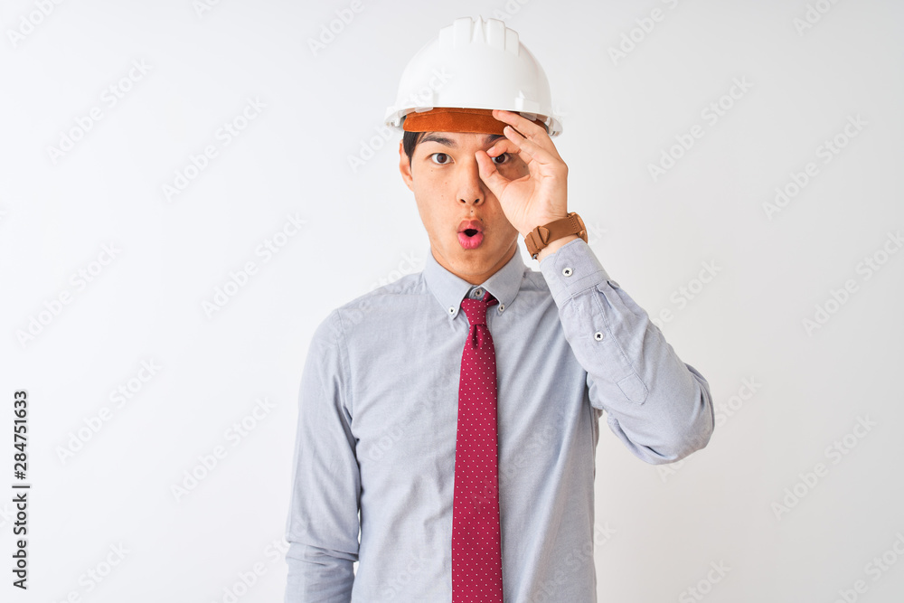 Chinese architect man wearing tie and helmet standing over isolated white background doing ok gesture shocked with surprised face, eye looking through fingers. Unbelieving expression. Chinese architect man wearing tie and helmet standing over isolated white background doing ok gesture shocked with surprised face, eye looking through fingers. Unbelieving expression.