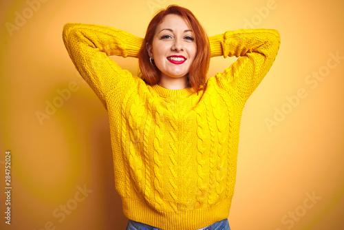 Beautiful redhead woman wearing winter sweater standing over isolated yellow background relaxing and stretching, arms and hands behind head and neck smiling happy © Krakenimages.com
