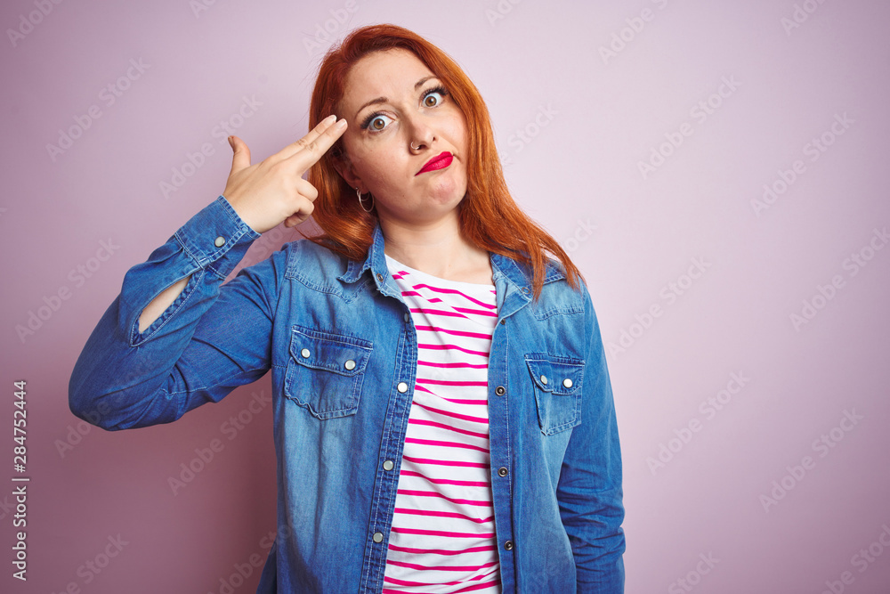 Beautiful redhead woman wearing denim shirt and striped t-shirt over isolated pink background Shooting and killing oneself pointing hand and fingers to head like gun, suicide gesture. Beautiful redhead woman wearing denim shirt and striped t-shirt over isolated pink background Shooting and killing oneself pointing hand and fingers to head like gun, suicide gesture.