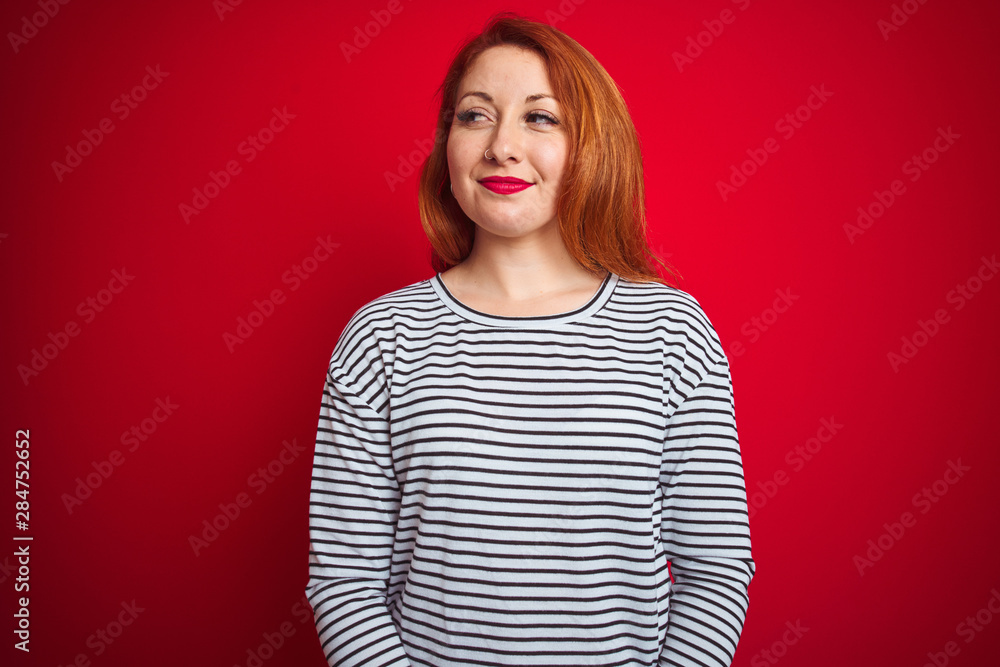 Young redhead woman wearing strapes navy shirt standing over red isolated background smiling looking to the side and staring away thinking. Young redhead woman wearing strapes navy shirt standing over red isolated background smiling looking to the side and staring away thinking.