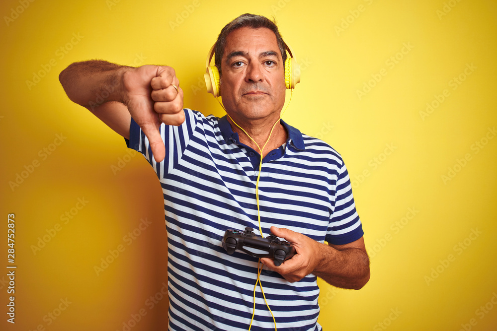 Middle age man playing video game using headphones over isolated yellow background with angry face, negative sign showing dislike with thumbs down, rejection concept Middle age man playing video game using headphones over isolated yellow background with angry face, negative sign showing dislike with thumbs down, rejection concept
