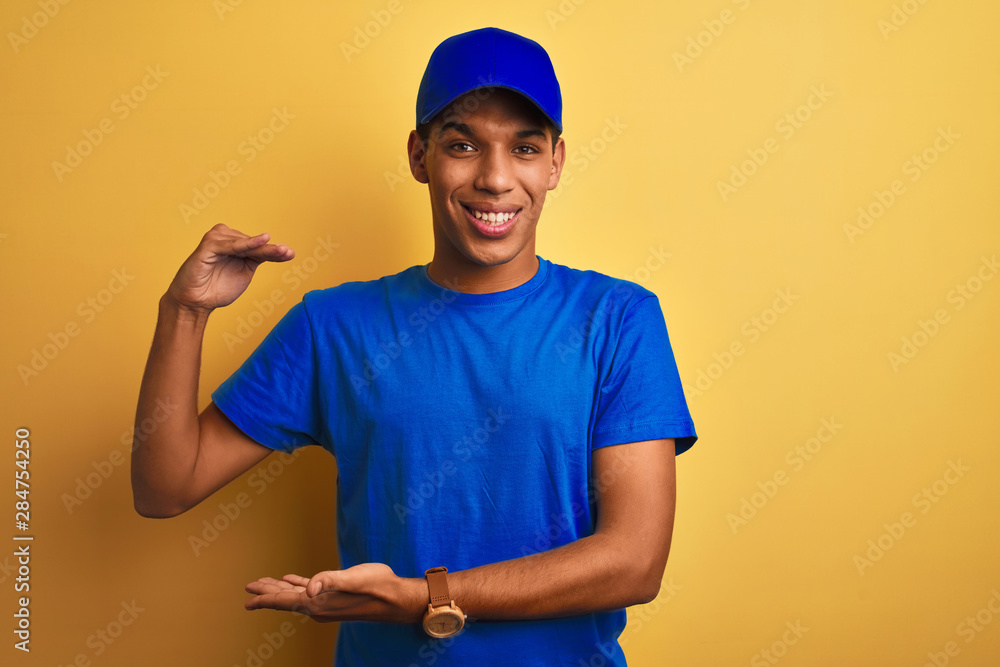 Young handsome arab delivery man standing over isolated yellow background gesturing with hands showing big and large size sign, measure symbol. Smiling looking at the camera. Measuring concept.