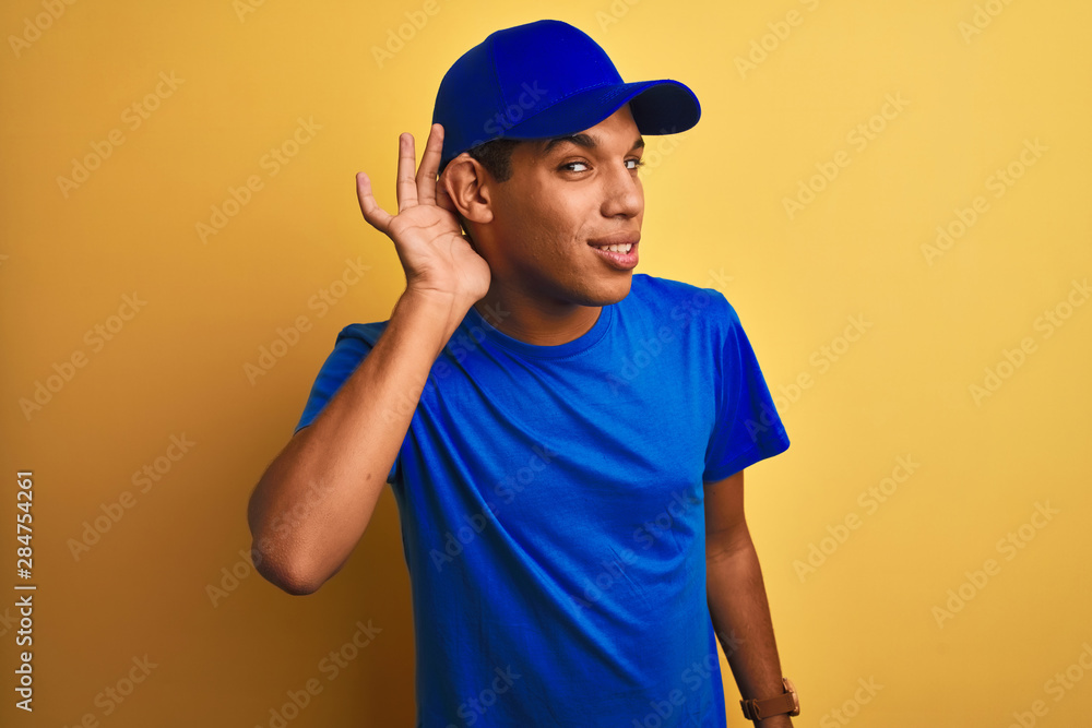 Young handsome arab delivery man standing over isolated yellow background smiling with hand over ear listening an hearing to rumor or gossip. Deafness concept.