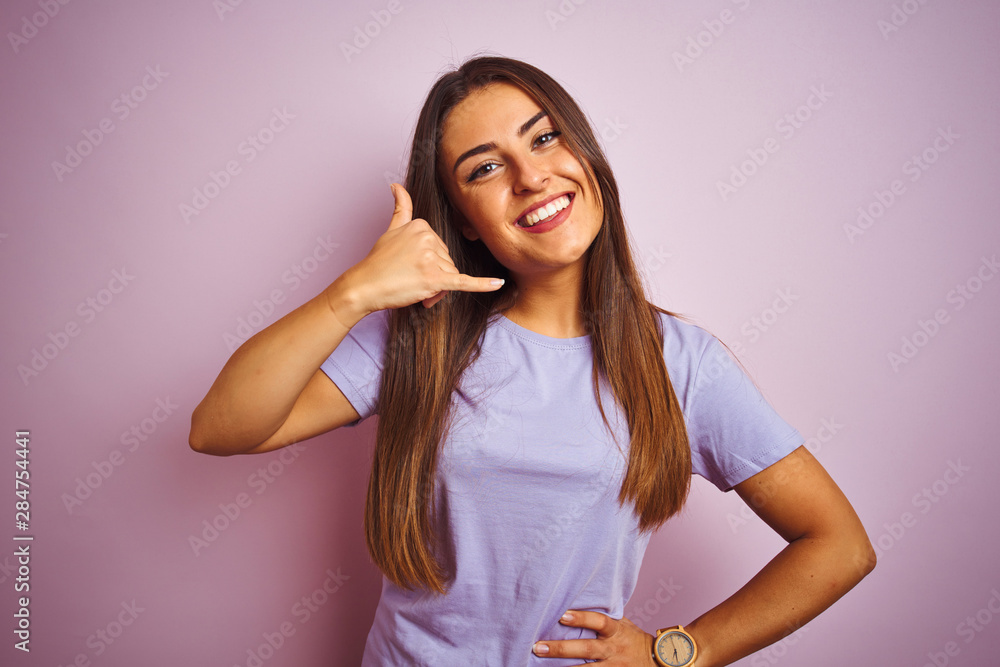 Fototapeta premium Young beautiful woman wearing casual t-shirt standing over isolated pink background smiling doing phone gesture with hand and fingers like talking on the telephone. Communicating concepts.