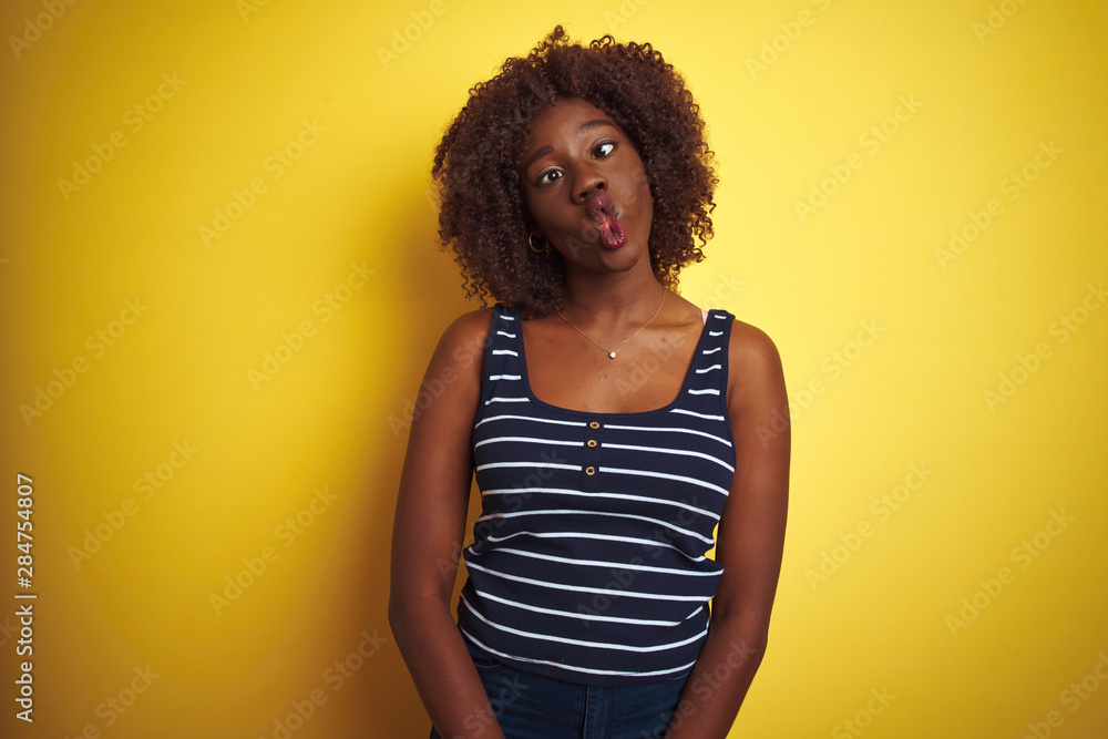 Young african afro woman wearing striped t-shirt over isolated yellow background making fish face with lips, crazy and comical gesture. Funny expression. Young african afro woman wearing striped t-shirt over isolated yellow background making fish face with lips, crazy and comical gesture. Funny expression.