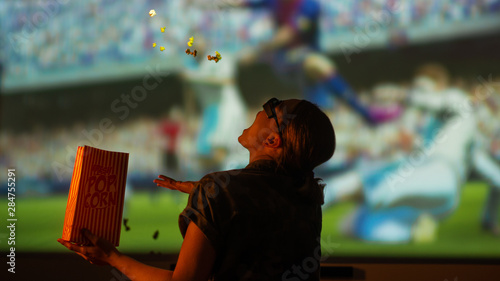 The girl is watching football, soccer in 3D,in glasses with popcorn frozen in the air, frozen in motion on a large screen.with entertainment and relaxation. action, rest in the cinema. Cheerleader,fan