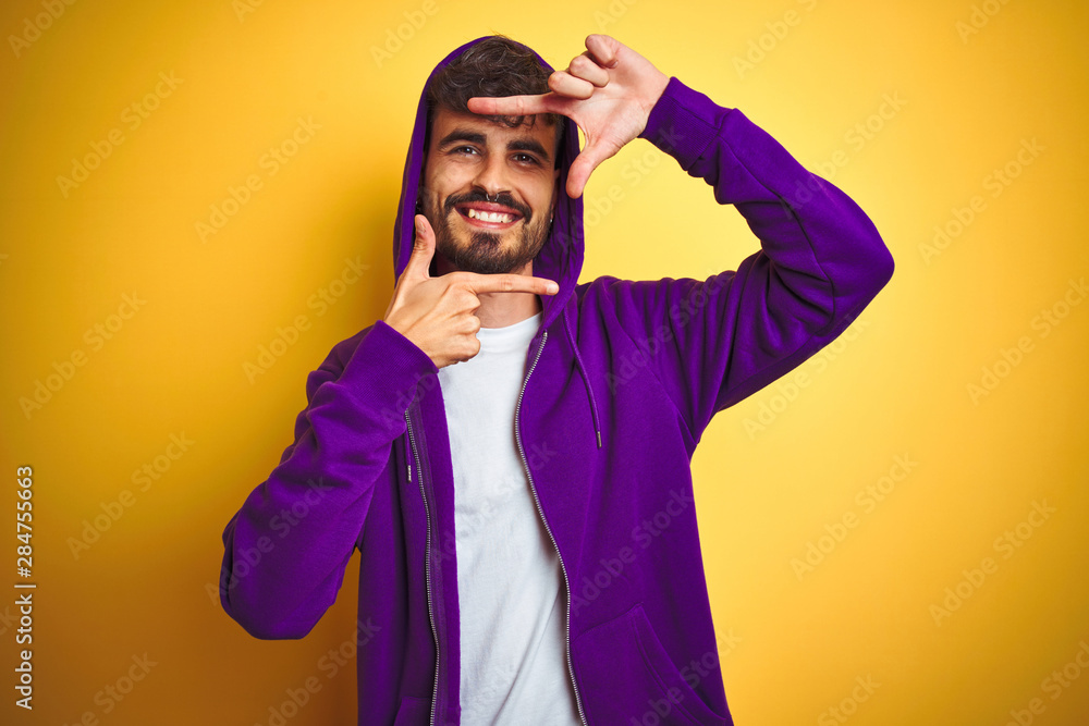 Young man with tattoo wearing purple sweatshirt with hood over isolated yellow background smiling making frame with hands and fingers with happy face. Creativity and photography concept. Young man with tattoo wearing purple sweatshirt with hood over isolated yellow background smiling making frame with hands and fingers with happy face. Creativity and photography concept.