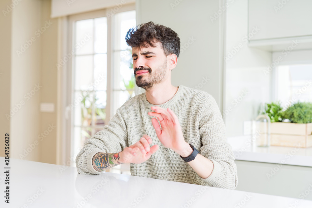 Young man wearing casual sweater sitting on white table disgusted expression, displeased and fearful doing disgust face because aversion reaction. With hands raised. Annoying concept.