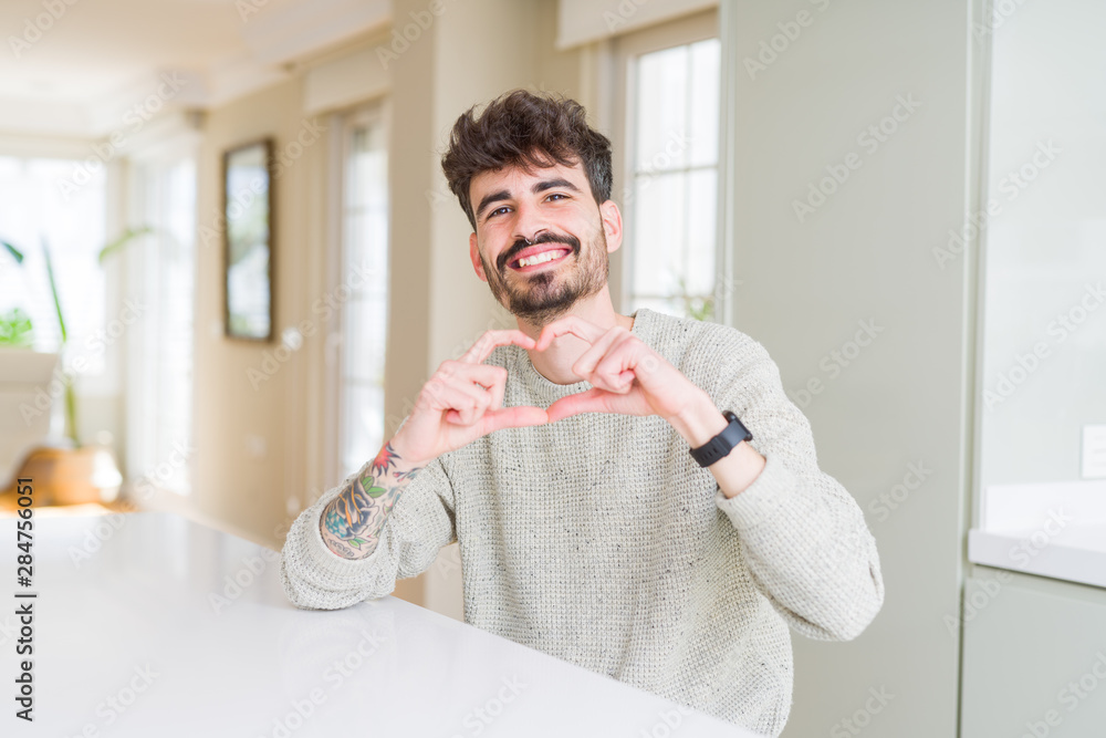 Young man wearing casual sweater sitting on white table smiling in love showing heart symbol and shape with hands. Romantic concept.
