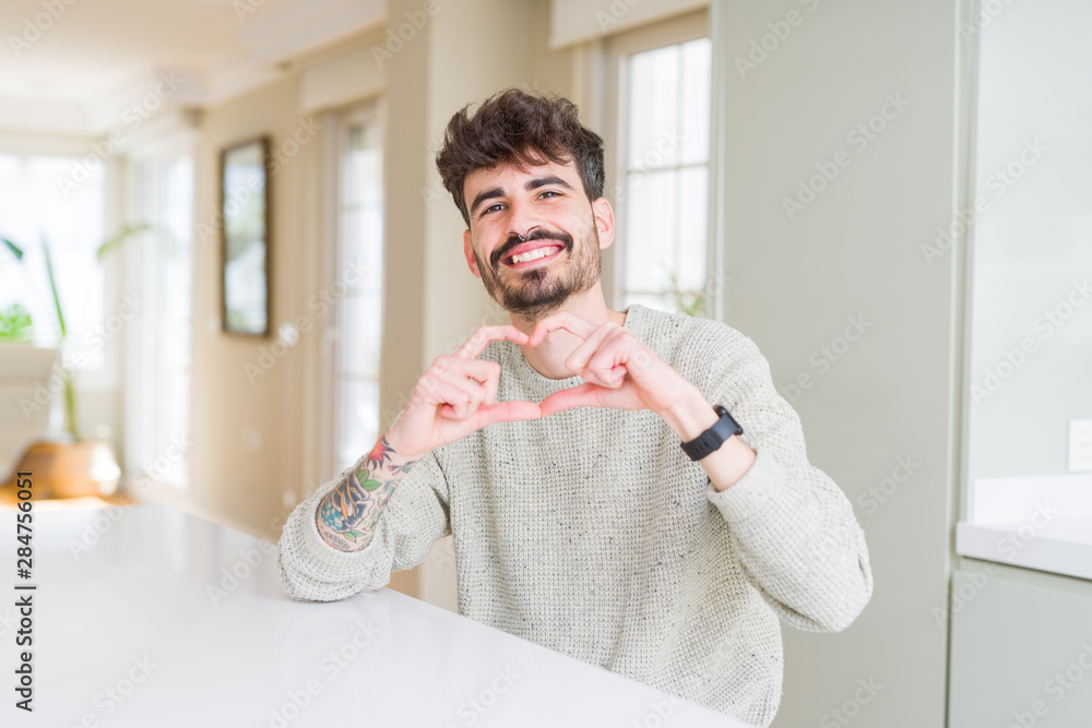 Fototapeta premium Young man wearing casual sweater sitting on white table smiling in love showing heart symbol and shape with hands. Romantic concept.
