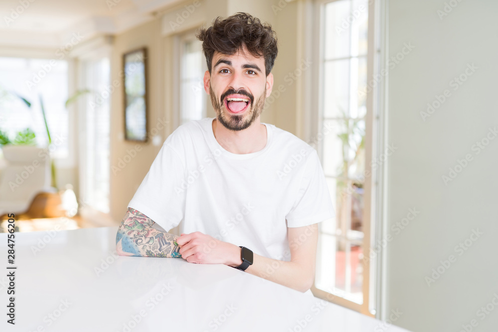 Young man wearing casual t-shirt sitting on white table sticking tongue out happy with funny expression. Emotion concept. Young man wearing casual t-shirt sitting on white table sticking tongue out happy with funny expression. Emotion concept.