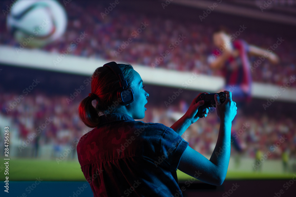 Fototapeta premium Soccer game, girl gamer playing a game in football headphones on a big screen, with bright light and a dark room. Gameplay, streaming, e-sports.