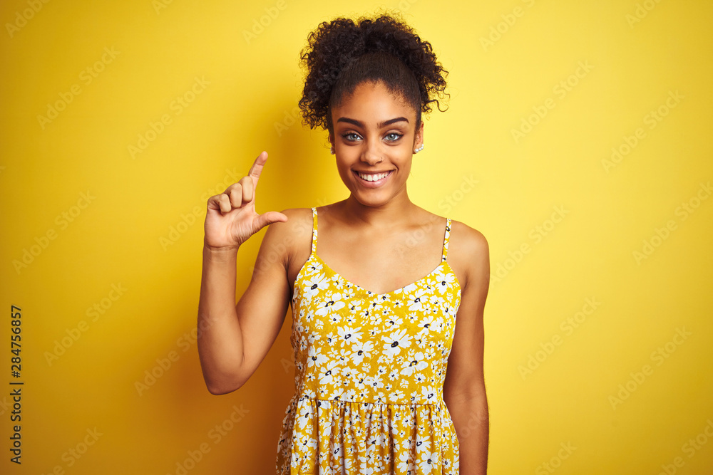 African american woman wearing casual floral dress standing over isolated yellow background smiling and confident gesturing with hand doing small size sign with fingers looking and the camera. African american woman wearing casual floral dress standing over isolated yellow background smiling and confident gesturing with hand doing small size sign with fingers looking and the camera.
