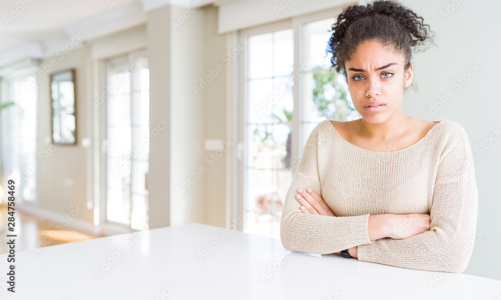 Beautiful young african american woman with afro hair sitting on table at home skeptic and nervous, disapproving expression on face with crossed arms. Negative person. Beautiful young african american woman with afro hair sitting on table at home skeptic and nervous, disapproving expression on face with crossed arms. Negative person.