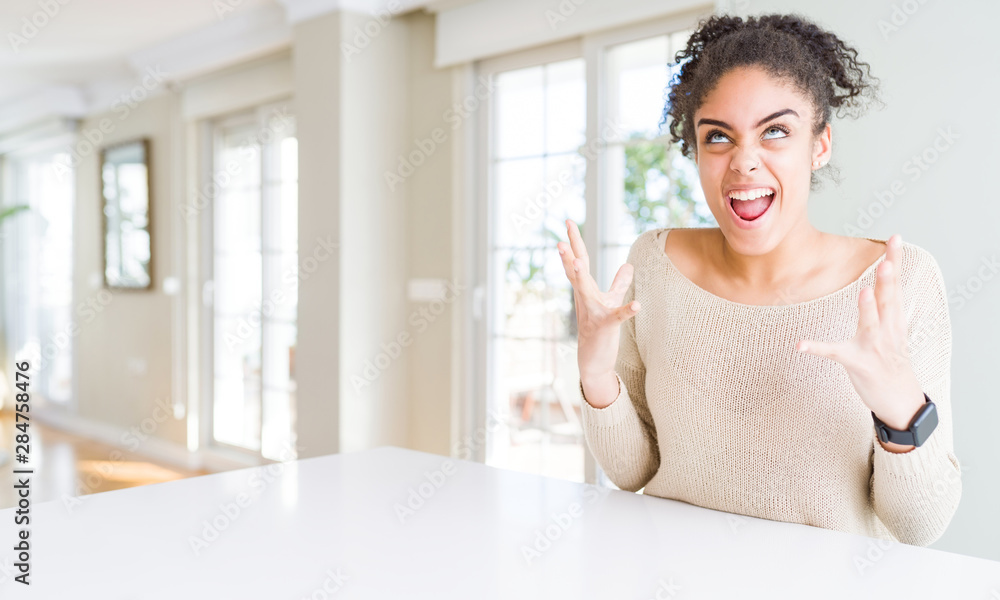 Beautiful young african american woman with afro hair sitting on table at home crazy and mad shouting and yelling with aggressive expression and arms raised. Frustration concept. Beautiful young african american woman with afro hair sitting on table at home crazy and mad shouting and yelling with aggressive expression and arms raised. Frustration concept.