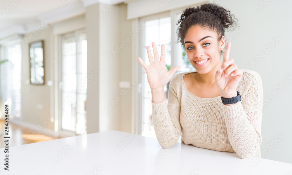 Beautiful young african american woman with afro hair sitting on table at home showing and pointing up with fingers number six while smiling confident and happy. Beautiful young african american woman with afro hair sitting on table at home showing and pointing up with fingers number six while smiling confident and happy.