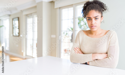 Beautiful young african american woman with afro hair sitting on table at home skeptic and nervous, disapproving expression on face with crossed arms. Negative person.