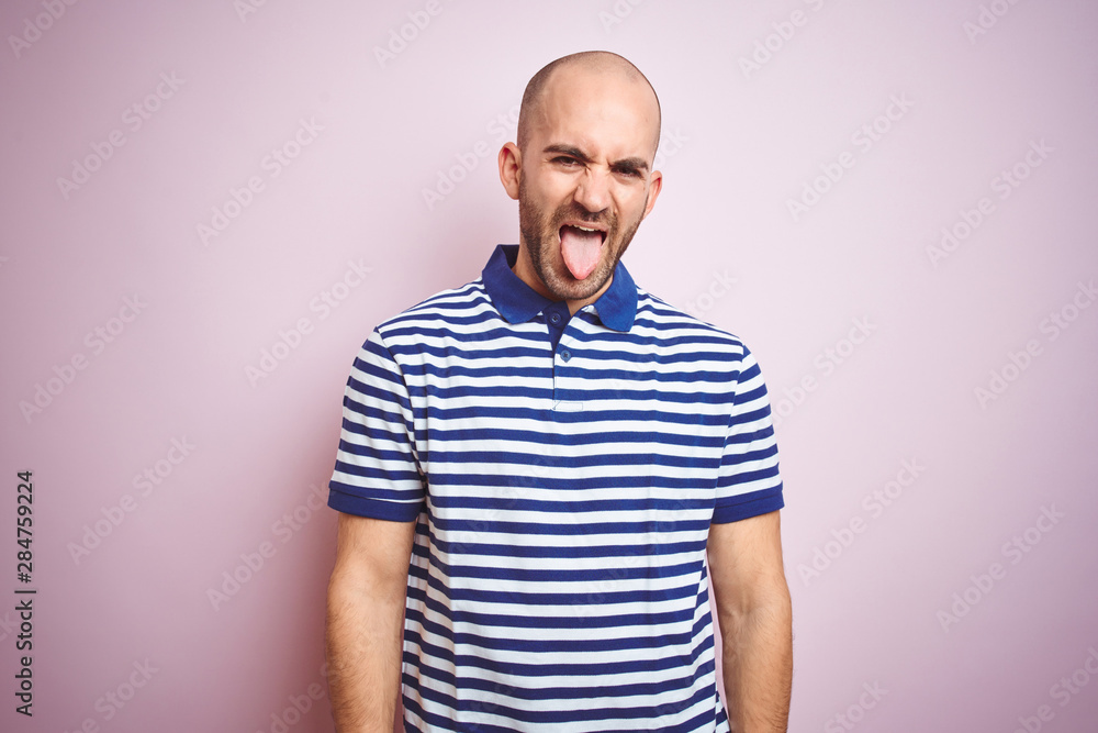 Obraz premium Young bald man with beard wearing casual striped blue t-shirt over pink isolated background sticking tongue out happy with funny expression. Emotion concept.