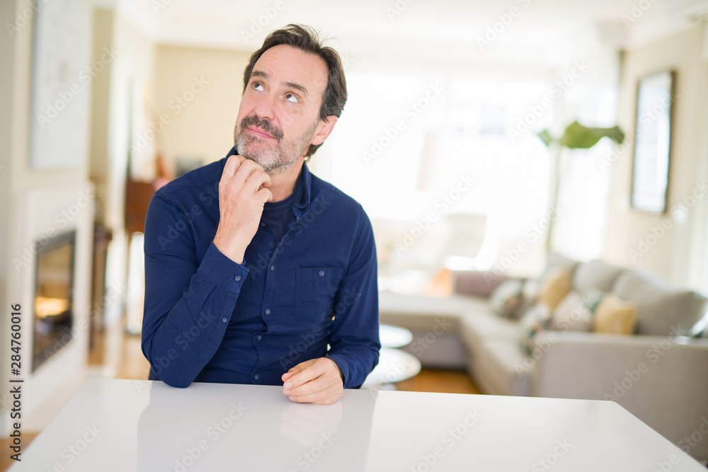 Handsome middle age man at home with hand on chin thinking about question, pensive expression. Smiling with thoughtful face. Doubt concept.
