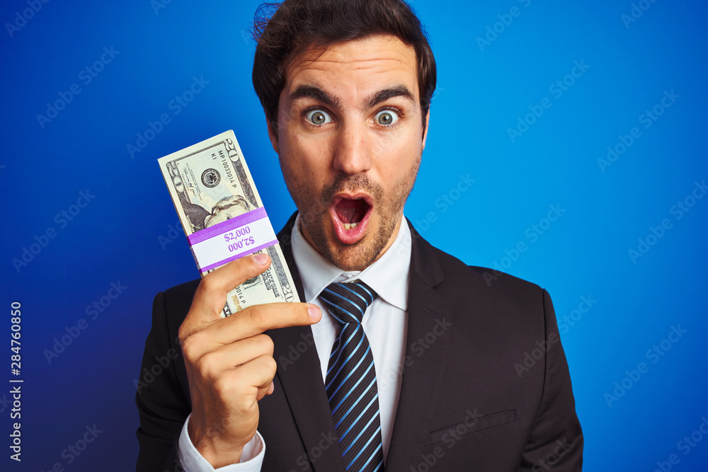 Young handsome businessman wearing suit holding dollars over isolated blue background scared in shock with a surprise face, afraid and excited with fear expression Young handsome businessman wearing suit holding dollars over isolated blue background scared in shock with a surprise face, afraid and excited with fear expression