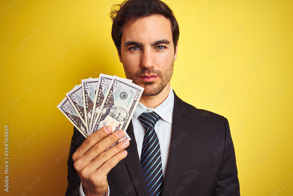 Young handsome businessman wearing suit holding dollars over isolated yellow background with a confident expression on smart face thinking serious Young handsome businessman wearing suit holding dollars over isolated yellow background with a confident expression on smart face thinking serious