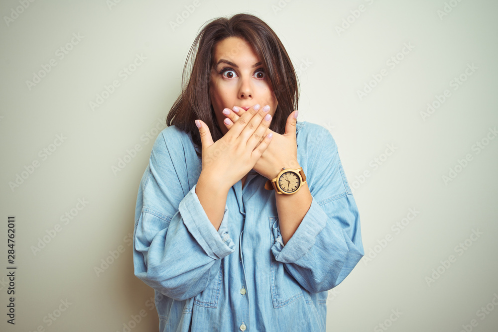 Young beautiful brunette woman wearing casual blue denim shirt over isolated background shocked covering mouth with hands for mistake. Secret concept. Young beautiful brunette woman wearing casual blue denim shirt over isolated background shocked covering mouth with hands for mistake. Secret concept.