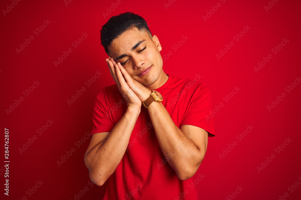 Young brazilian man wearing t-shirt standing over isolated red background sleeping tired dreaming and posing with hands together while smiling with closed eyes. Young brazilian man wearing t-shirt standing over isolated red background sleeping tired dreaming and posing with hands together while smiling with closed eyes.