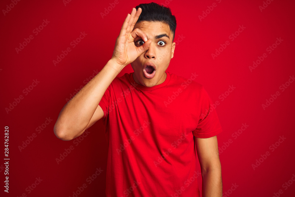 Young brazilian man wearing t-shirt standing over isolated red background doing ok gesture shocked with surprised face, eye looking through fingers. Unbelieving expression. Young brazilian man wearing t-shirt standing over isolated red background doing ok gesture shocked with surprised face, eye looking through fingers. Unbelieving expression.