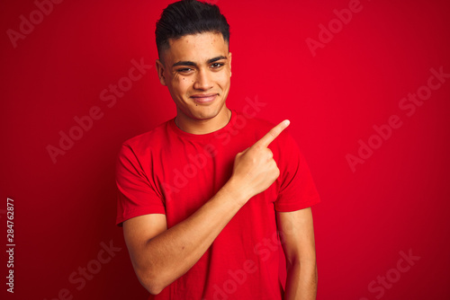 Young brazilian man wearing t-shirt standing over isolated red background Pointing with hand finger to the side showing advertisement, serious and calm face