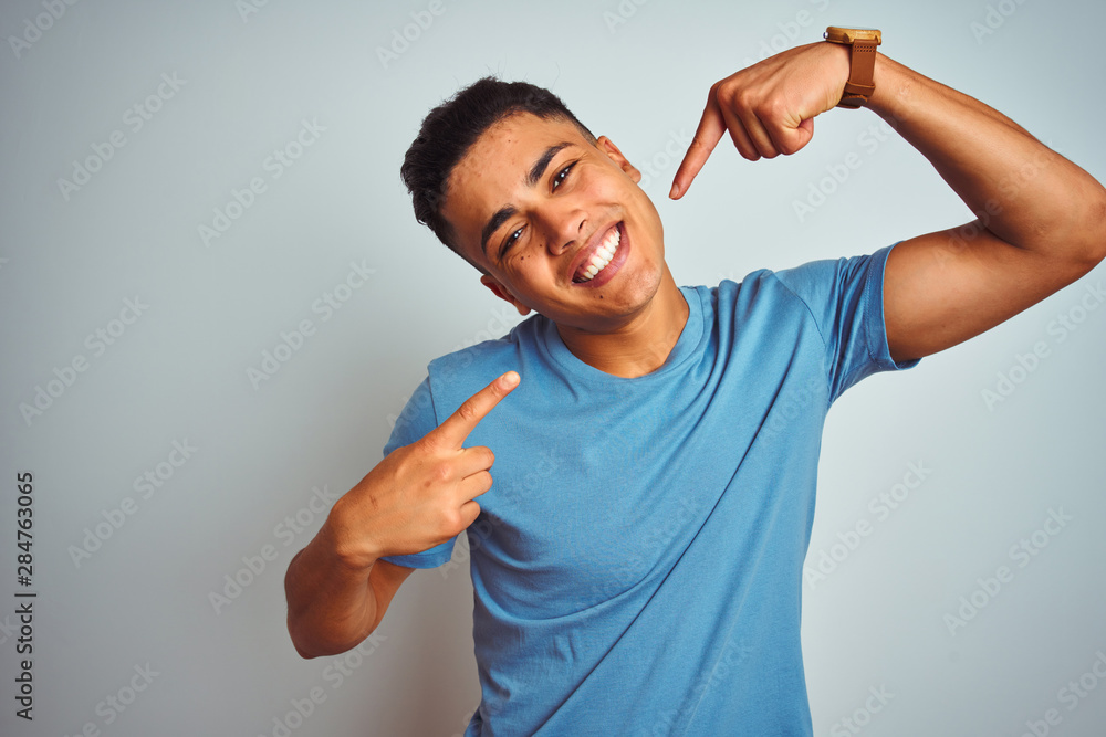 Fototapeta premium Young brazilian man wearing blue t-shirt standing over isolated white background smiling cheerful showing and pointing with fingers teeth and mouth. Dental health concept.