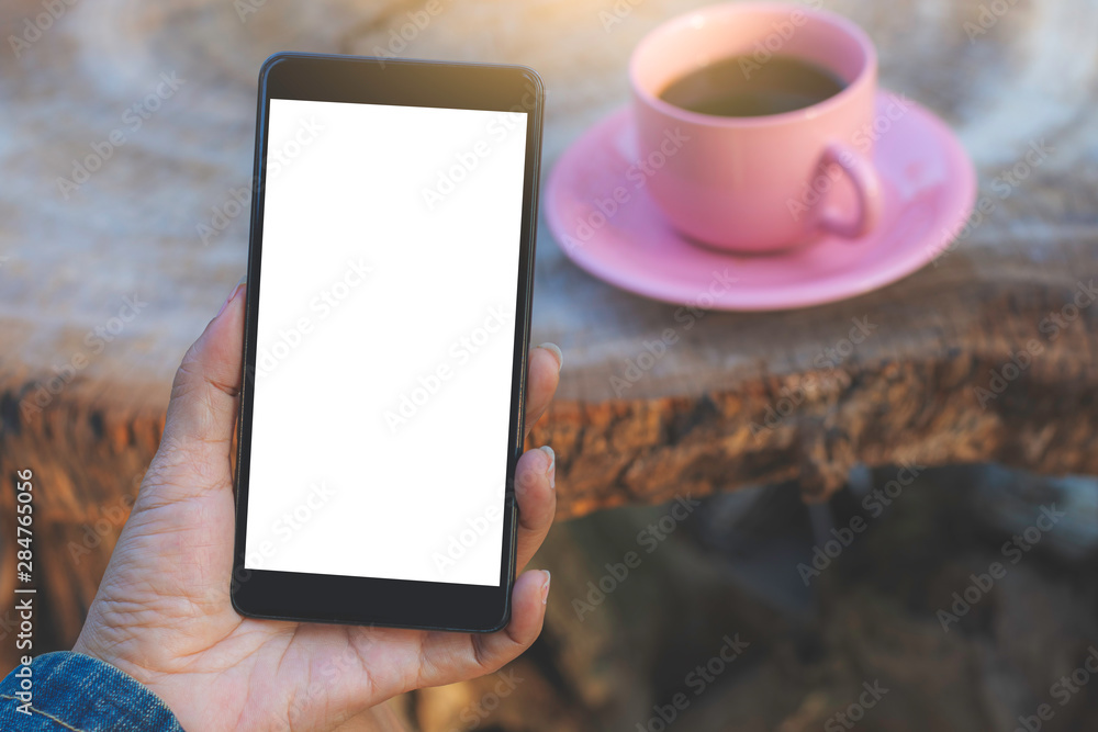 Desktop mobile mockup concept.Woman hand holding and showing mobile smart phone with blank white screen with pink cup of hot coffee on old wooden table at cafe outdoor. Vintage style.