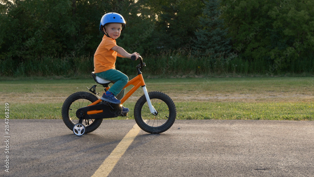 Obraz premium Cute little boy ( two years old child) is riding bike. Kid in orange t-shirt and blue helmet. Orange kids bicycle. First experience, childhood memories