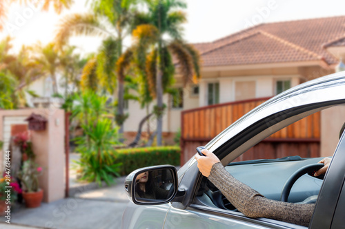 Woman in car, hand using remote control to open auto wooden door with modern home blurred background. Automatic gate concept